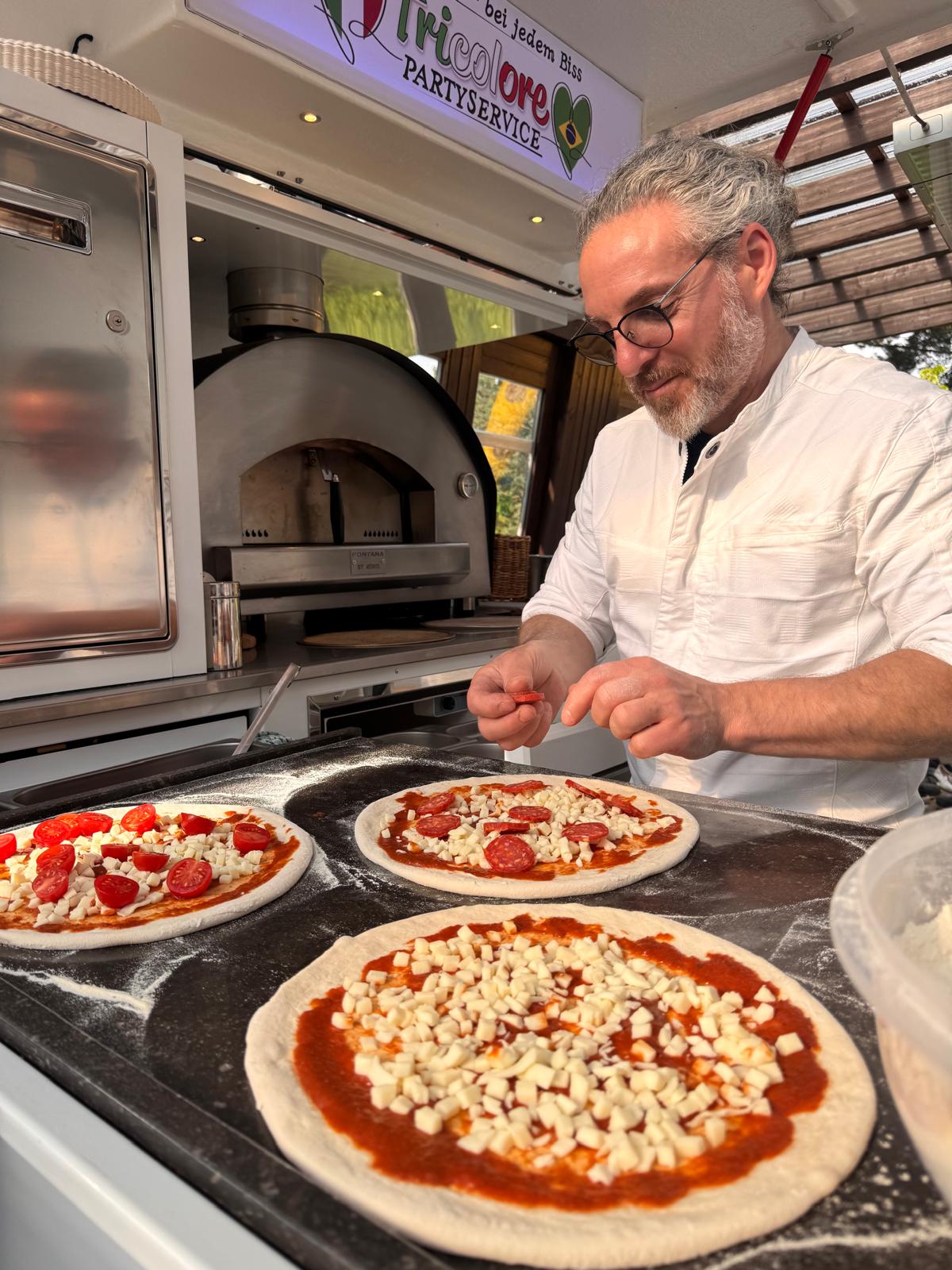 Giuseppe belegt frische Pizzen im Pizza Ape, Leuchtschild Tricolore Partyservice im Hintergrund